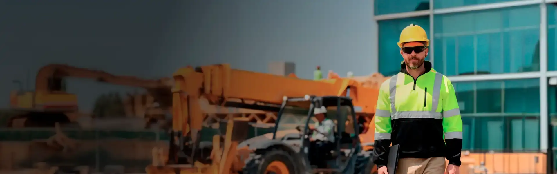 Worker in a hard hat and vest with a laptop near construction machinery